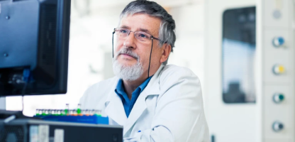 Male Scientist with white beard and salt and pepper hair and  glasses looking at monitor