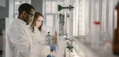 Two scientists wearing white lab coats in a brightly lit lab working on a science project. 