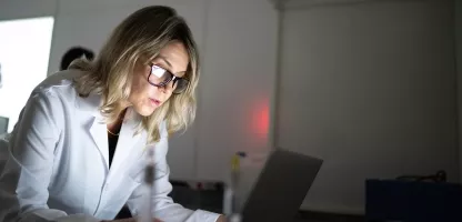 Woman in a white coat and glasses looking at a computer in a dimly lit room