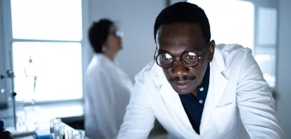 A man wearing glasses in a white coat looking at a computer screen in a scientific lab
