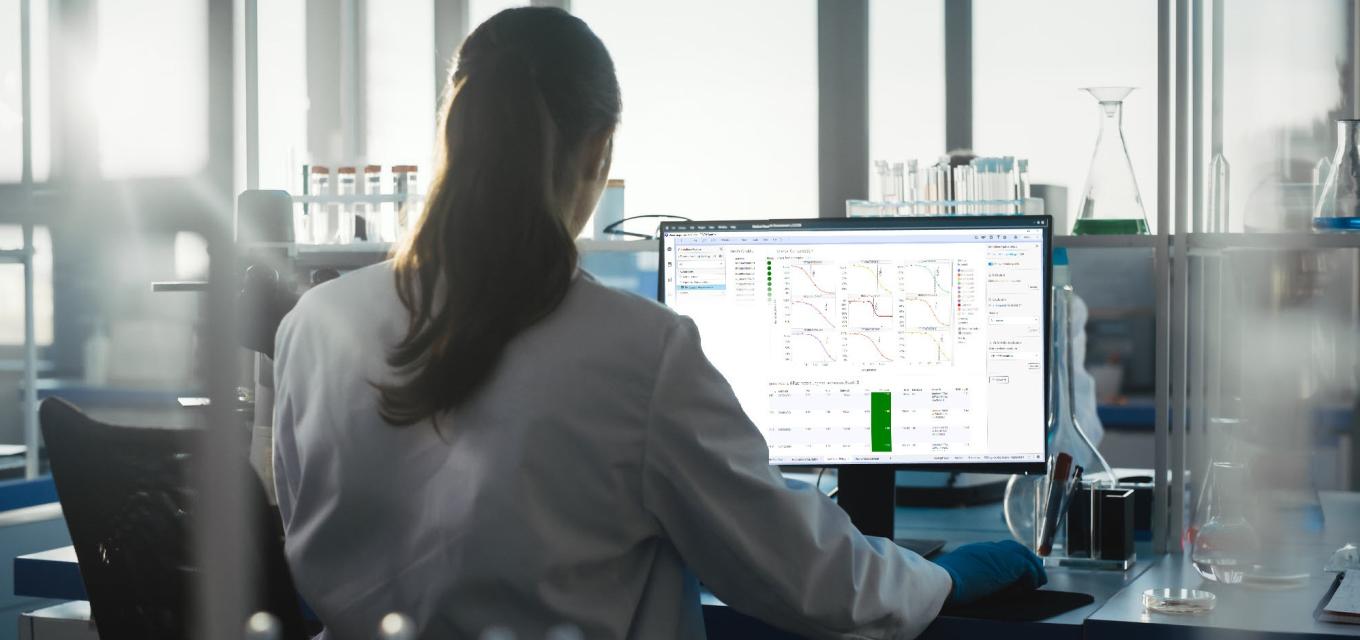 woman looking at monitor w test tubes SRS summit.png woman back with pony tail, looking at monitor in lab setting